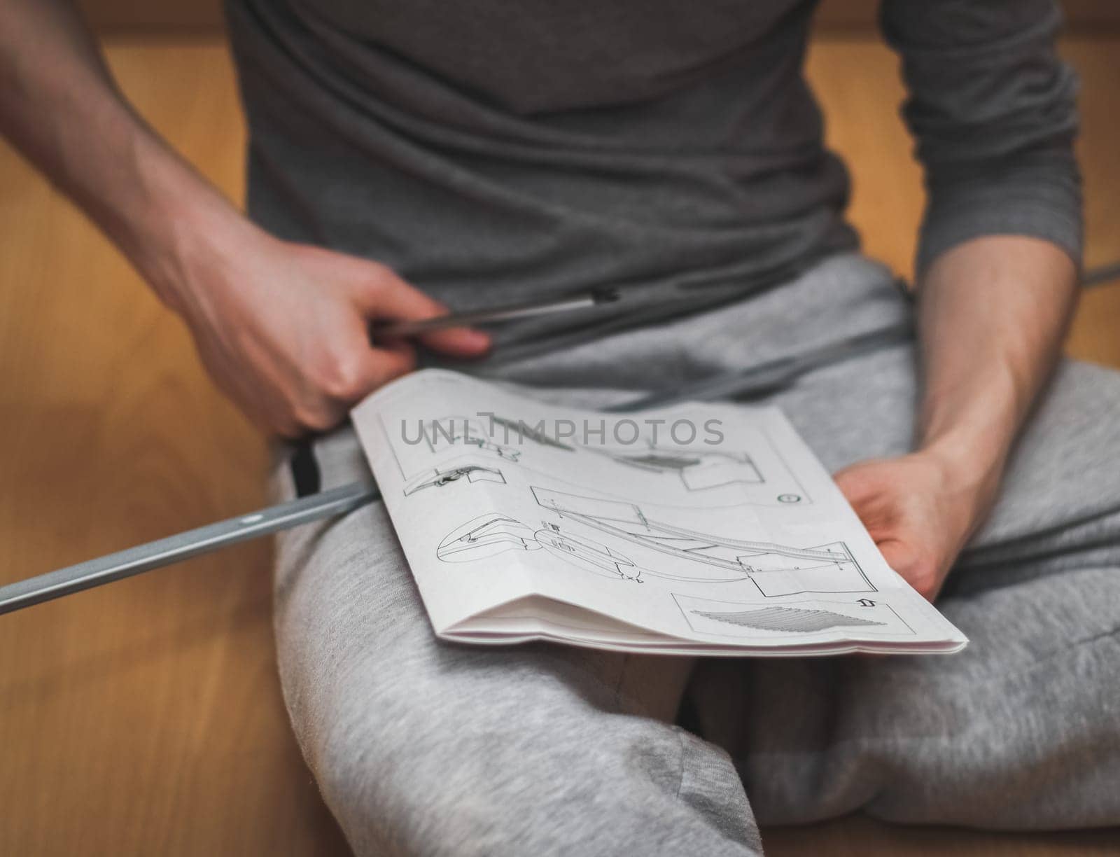 Caucasian young man in gray sweatpants and long-sleeved T-shirt reads book instructions for assembling a bed, sitting on the floor in his room with a screwdriver and a screw in his hands, close-up side view. Concept of assembling furniture, shopping, at home.