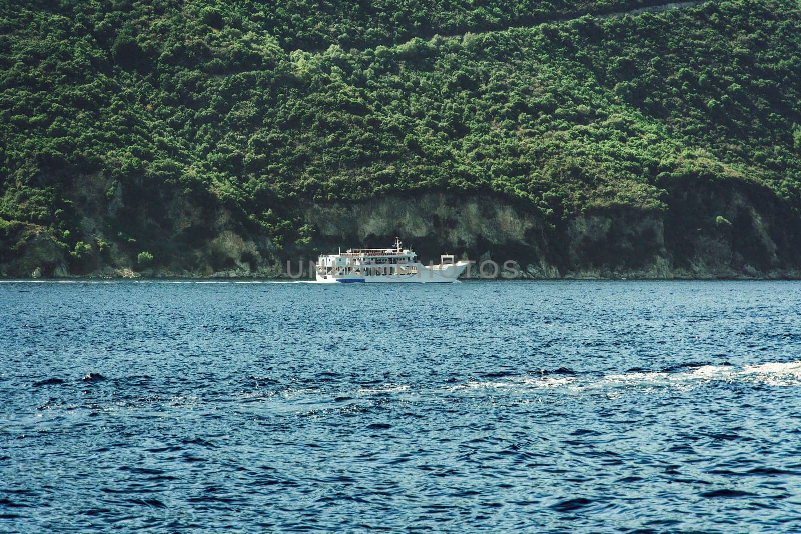 Small car and passengers ferry sailing next to a green plantation coastline in Lefkada Ionian Island Greece.