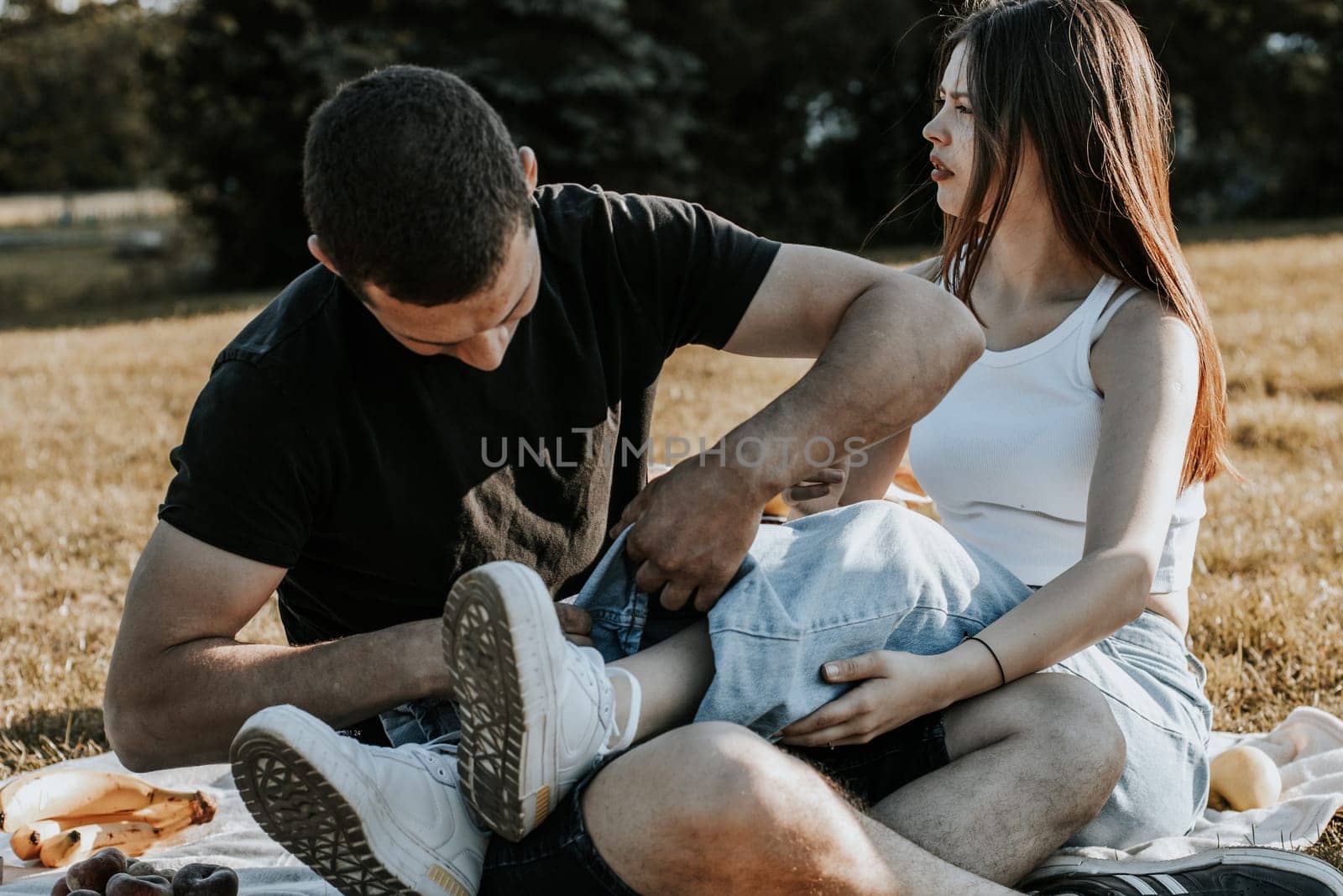 One beautiful Caucasian young brunette guy rolls up the hem of his girlfriend's jeans while sitting on a blanket with fruits in the park on a picnic against the backdrop of nature, close-up side view.