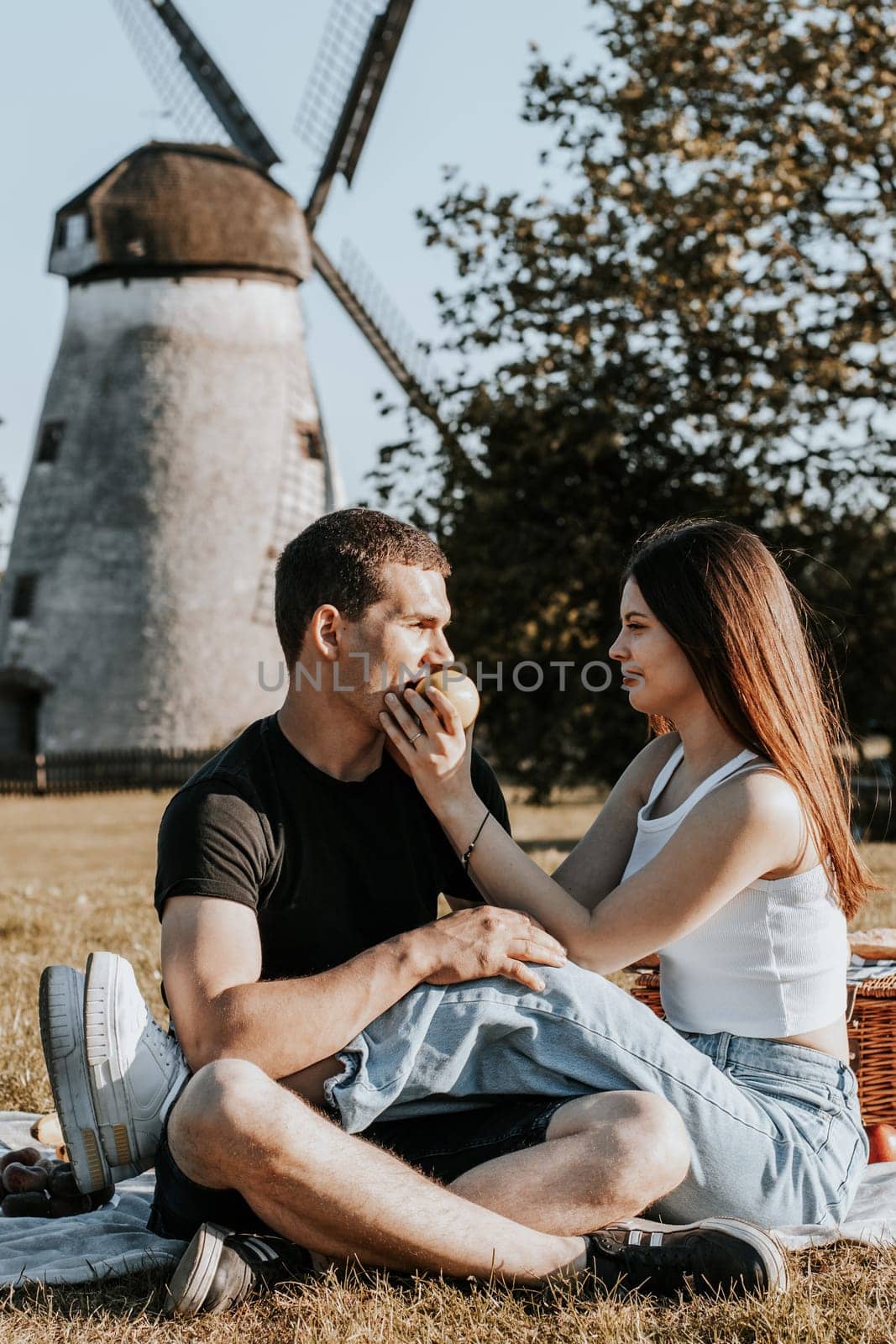 One beautiful Caucasian brunette girl brings an apple to her boyfriend s mouth wanting to treat him, sitting romantically sideways on a blanket with a basket in the park on a picnic against the backdrop of an old mill, close-up side view.