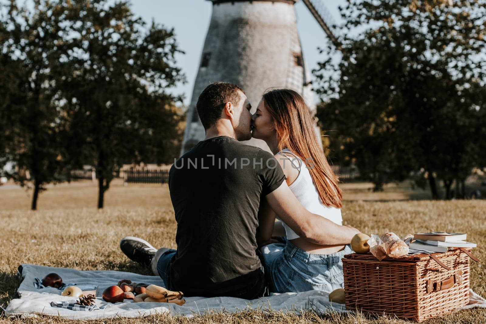 One beautiful Caucasian guy hugging a girl and gently kissing her on the lips on a sunny spring day, sitting with their backs on a blanket with a basket, books and fruits in the park on a picnic against the backdrop of an old mill, close-up view from below.