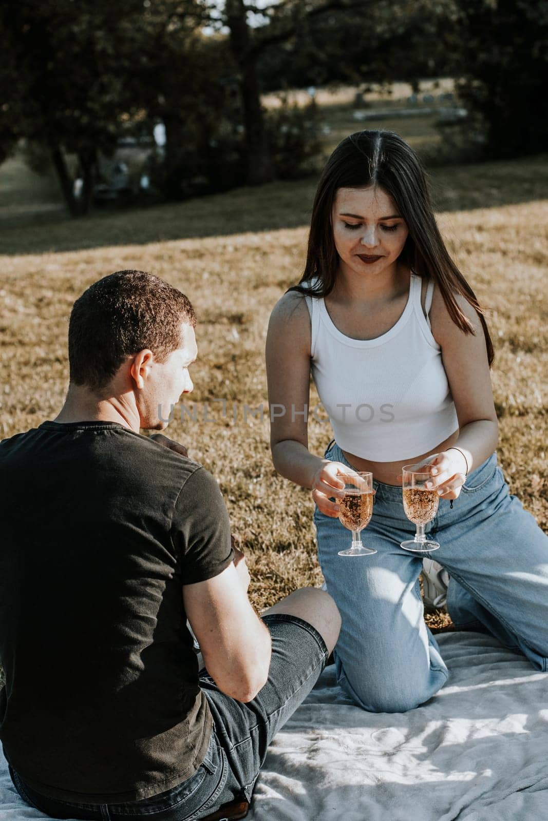 One beautiful Caucasian brunette girl with long flowing hair with a smile holds glasses of champagne in her hands and serves them to a guy sitting with his back turned in the park on a picnic, close-up side view.