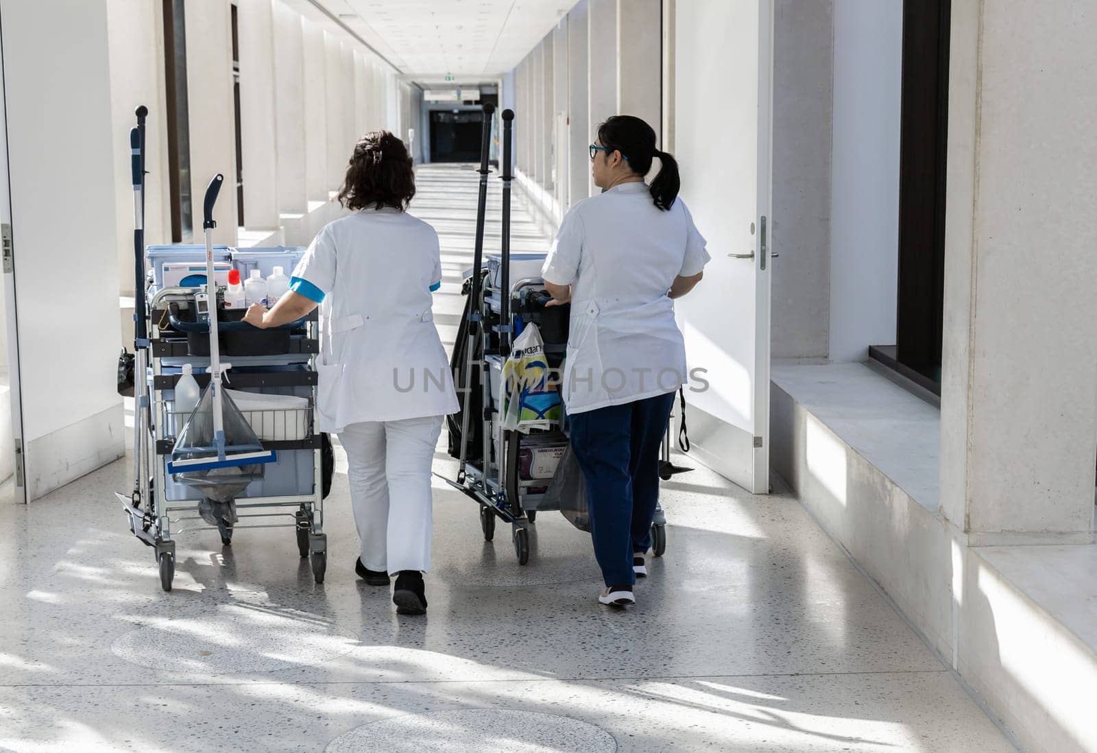 Two female cleaners with cleaning carts walk along a long white corridor. Cleaning concept in office and hospital.