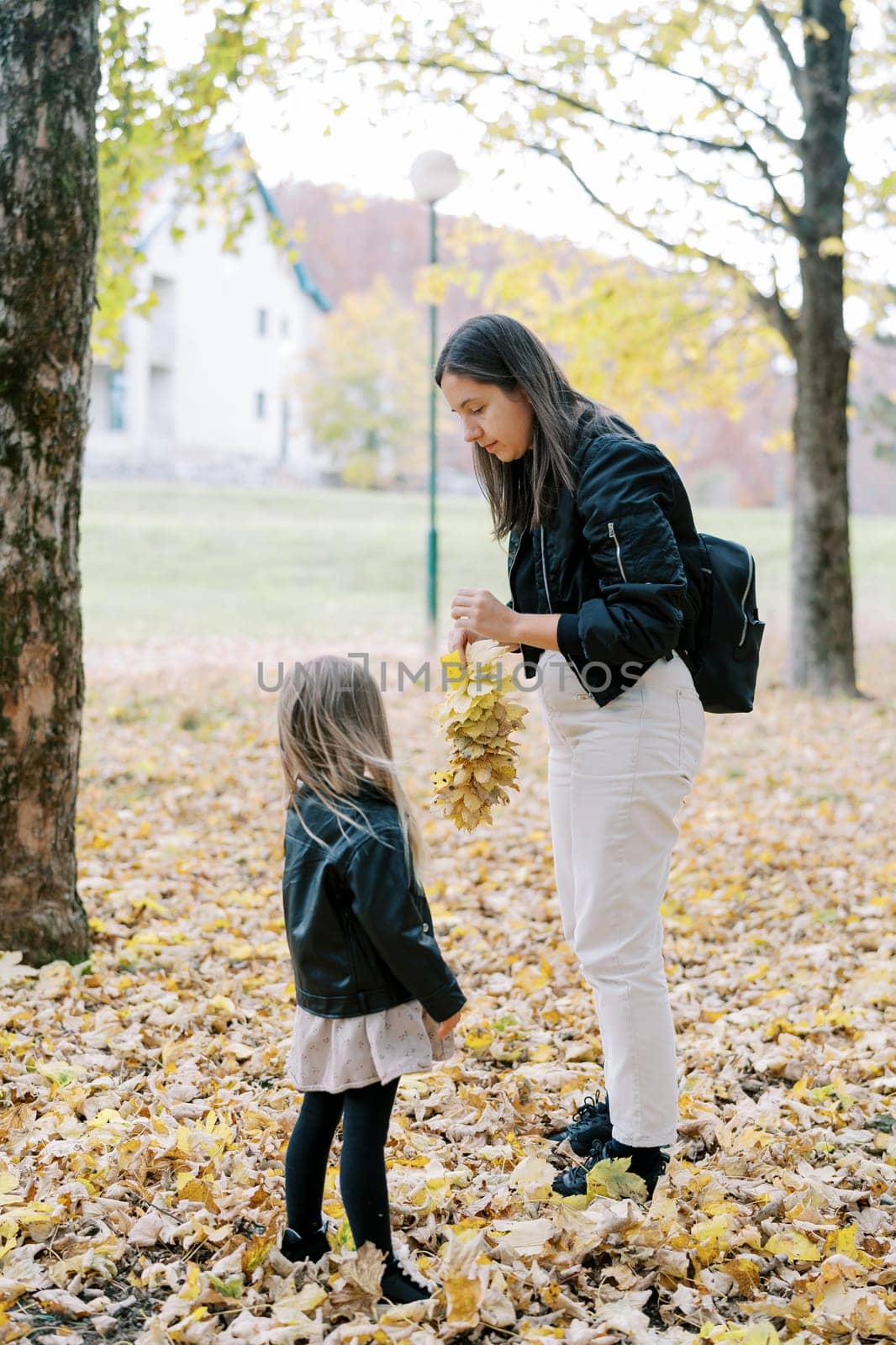 Little girl looks at her mother weaving a wreath of yellow autumn leaves while standing in the park. Back view. High quality photo