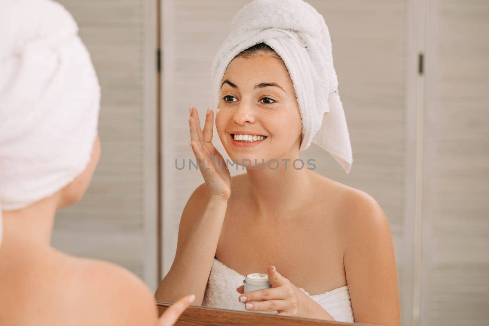 Portrait of beautiful woman caring of her skin. girl looking at mirror and applying cream on her face in front of the mirror in bathroom