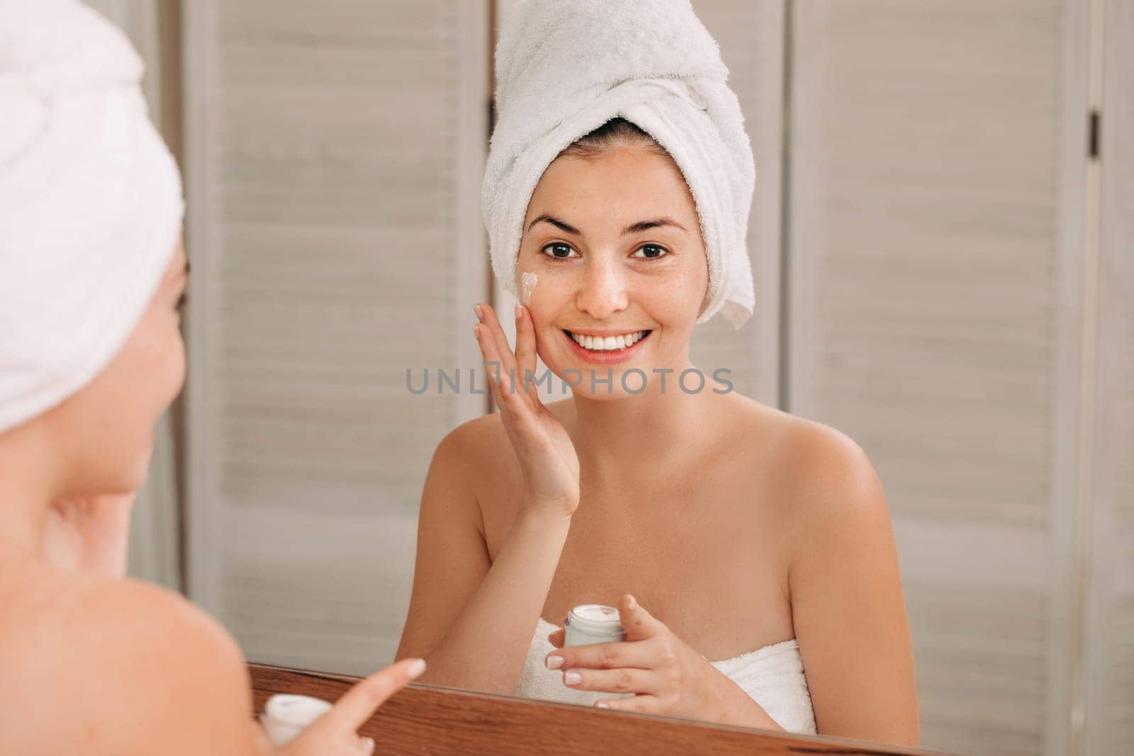 Portrait of beautiful woman caring of her skin. girl looking at mirror and applying cream on her face in front of the mirror in bathroom
