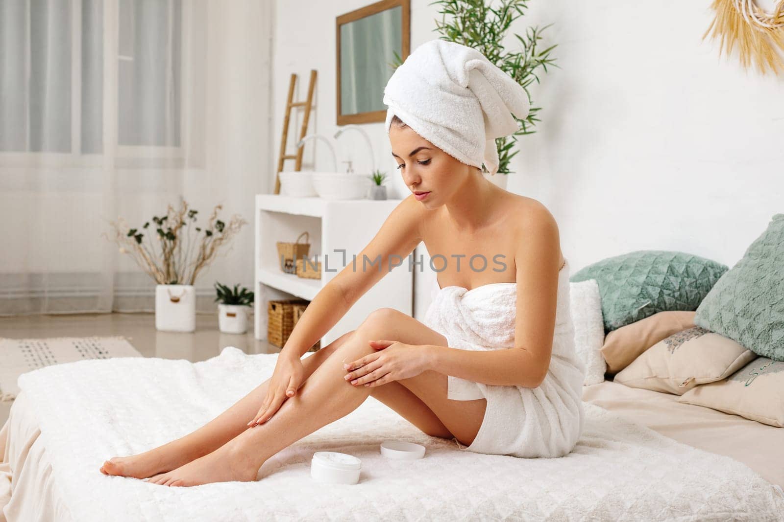 Young beautiful woman in towel applying cream on her legs after shower at home