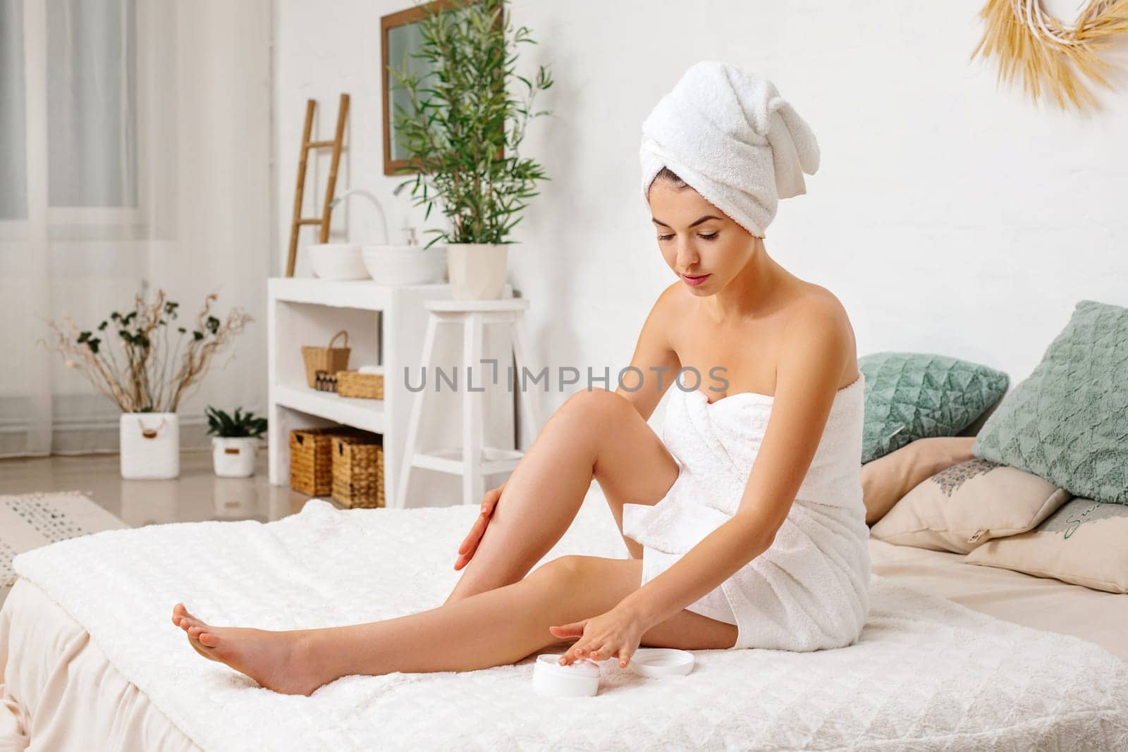 Young beautiful woman in towel applying body cream on her leg at home