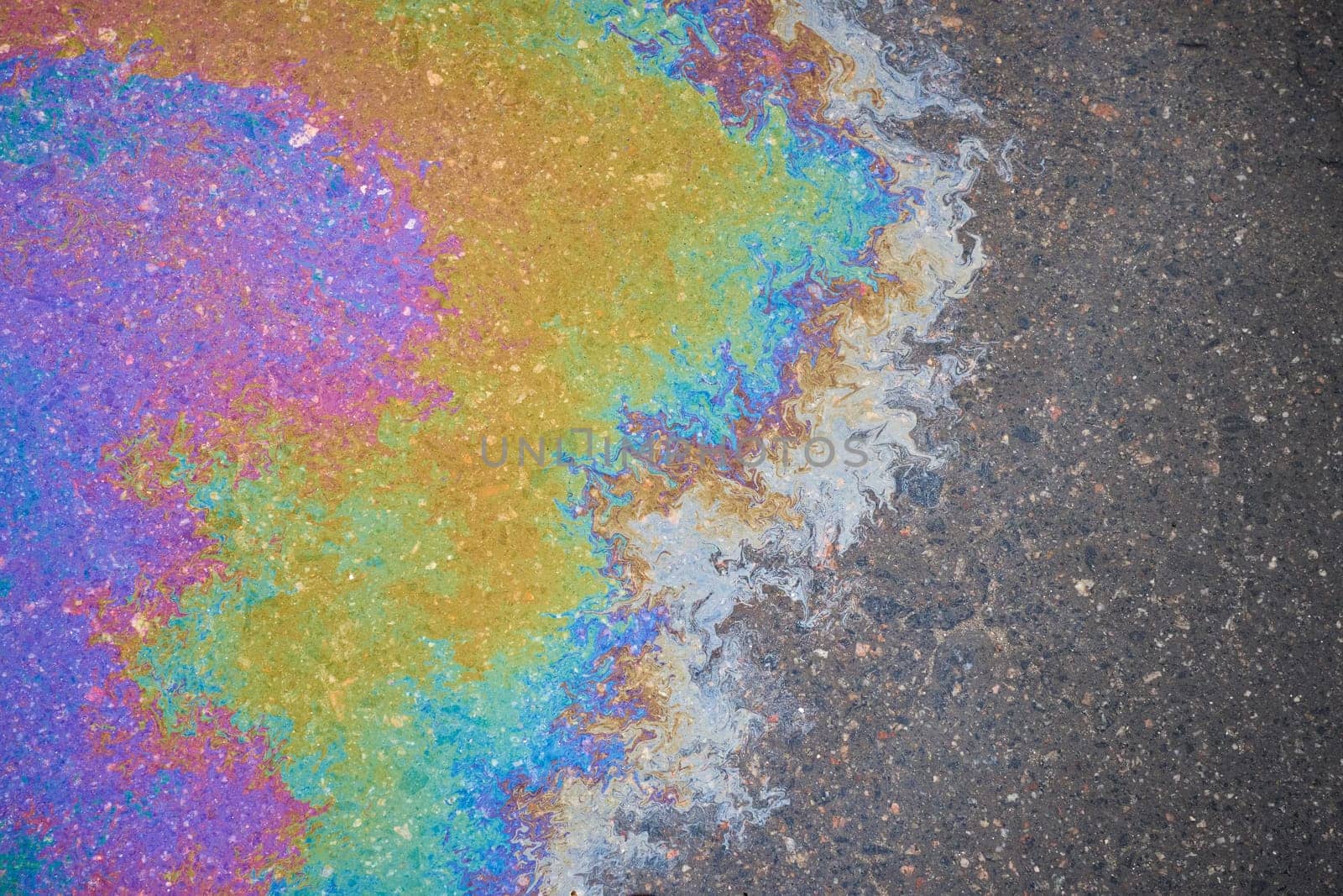 Residues of oil on asphalt left by rain form trails that scatter sunlight, creating a rainbow effect