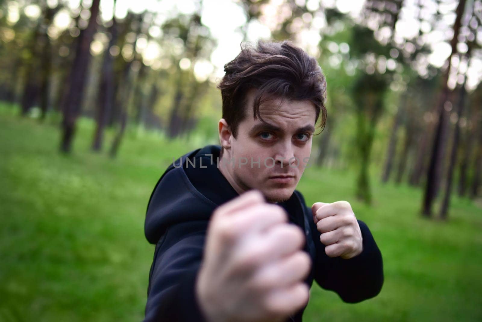sporty young man in a sporty black jacket in a fighting pose raising his fist for a punch against a background of forest trees.
