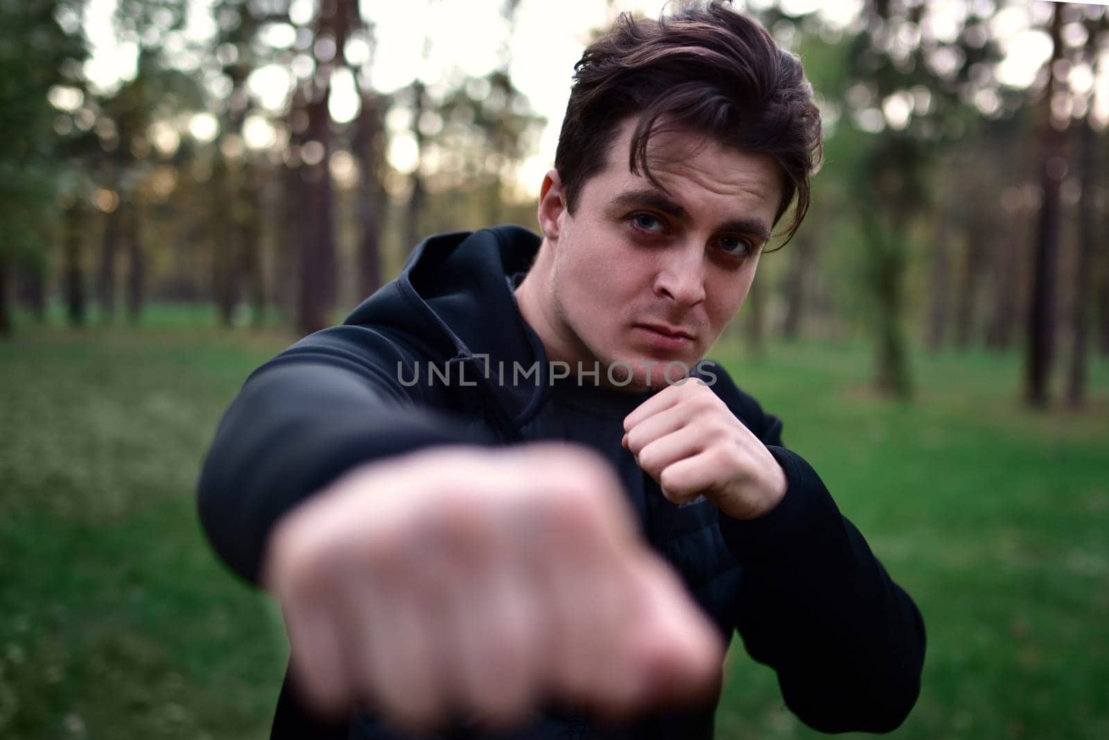 sporty young man in a sporty black jacket in a fighting pose raising his fist for a punch against a background of forest trees