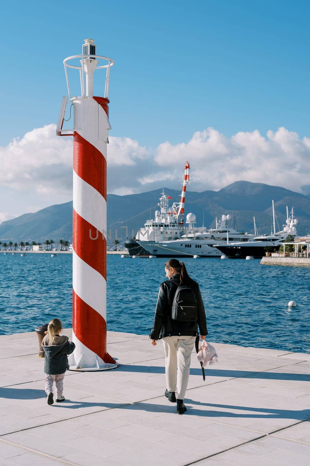 Mother and a little girl walk along the pier to the striped lighthouse. Back view. High quality photo