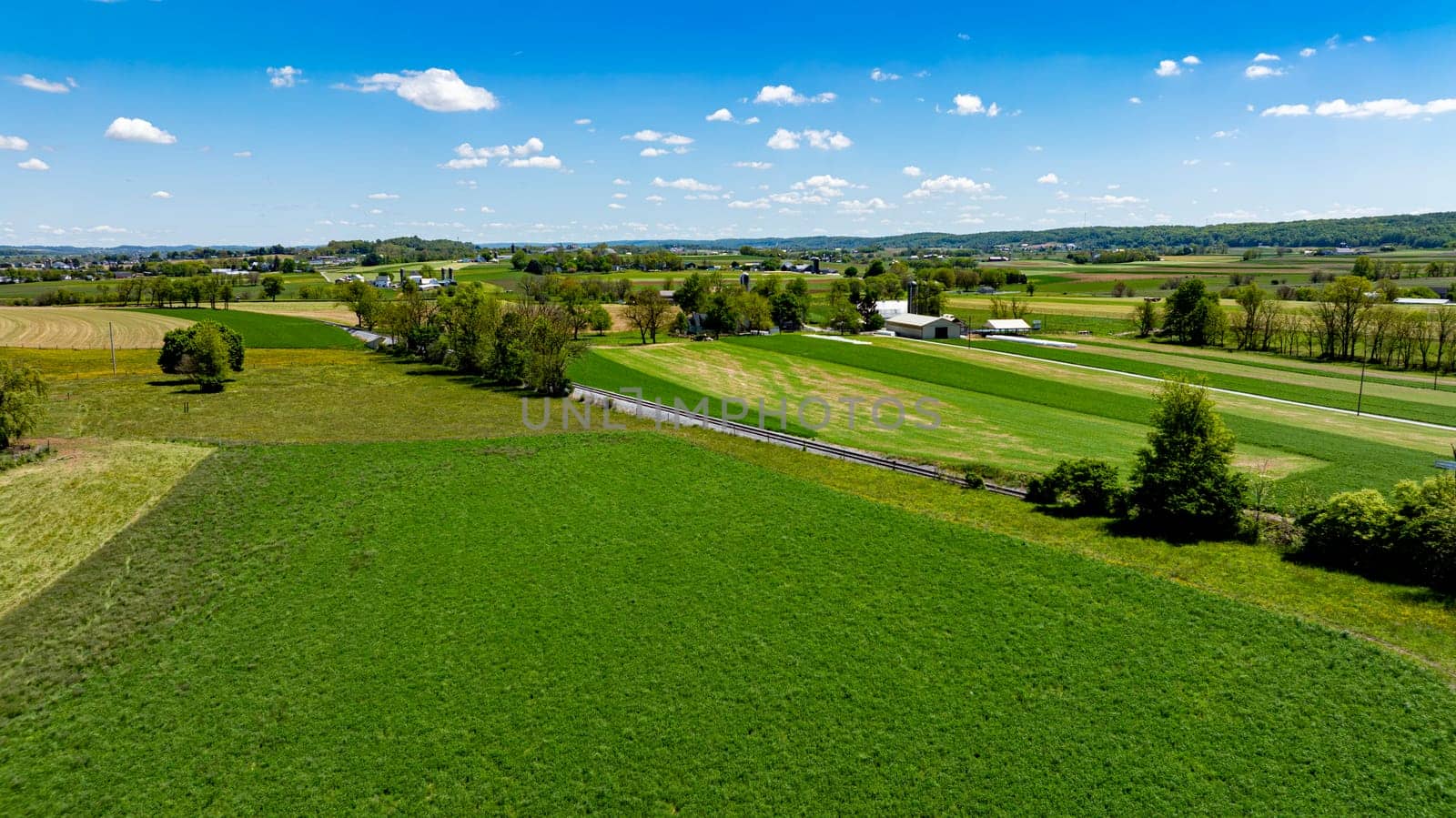 Stunning aerial view capturing a diverse agricultural landscape with fields of varying shades of green, a small road, and distant buildings under a sky dotted with clouds.