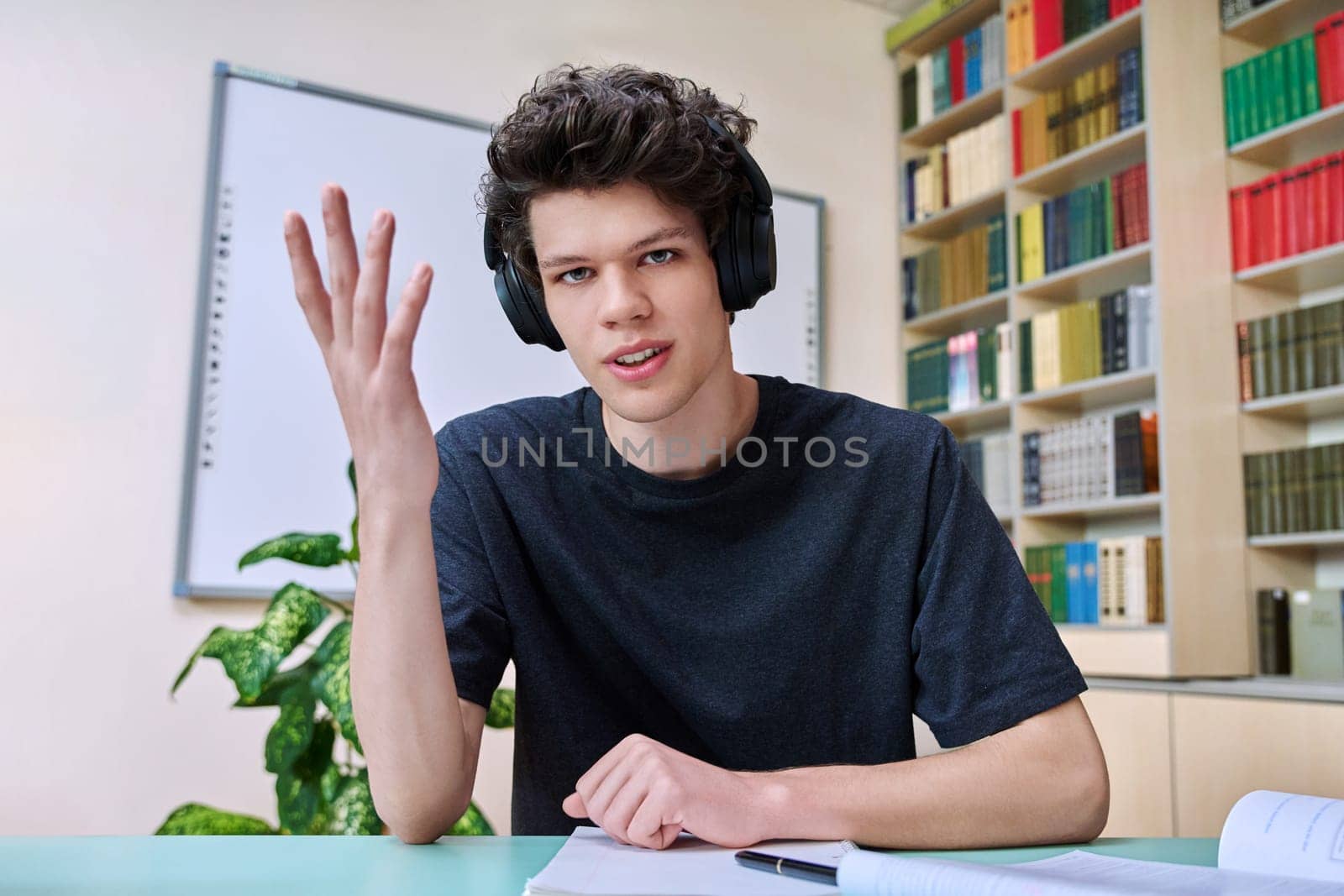 Web cam portrait of college student guy in headphones looking talking to camera in educational building library classroom. Video call chat conference, online lesson exam test, technology education