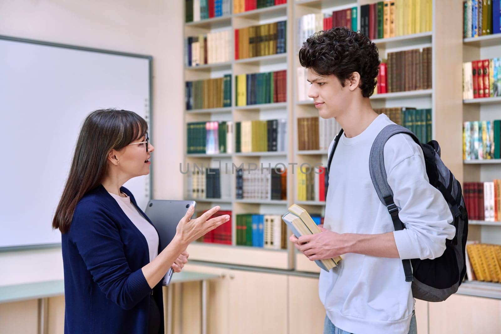 Female teacher talking to guy teenage college student inside educational library office. Education, teaching, youth, lifestyle concept