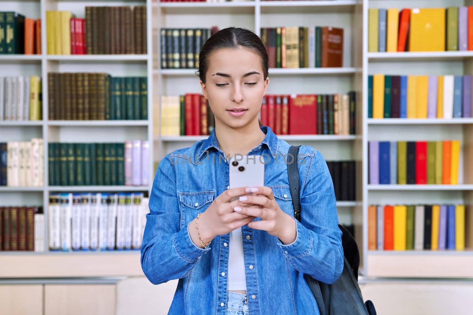 Teenage female student with backpack using smartphone, inside high school building, in library. Technologies, mobile educational apps applications, services, e-learning concept