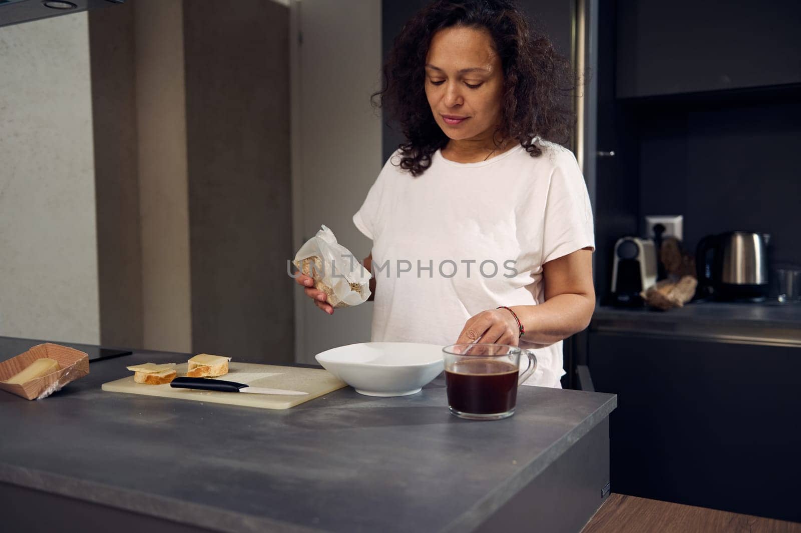 Beautiful happy mixed race woman in pajamas in the morning, pouring oat flakes into a white bowl, preparing dry breakfast, standing at kitchen counter at home. Food and drink concept. Healthy eating