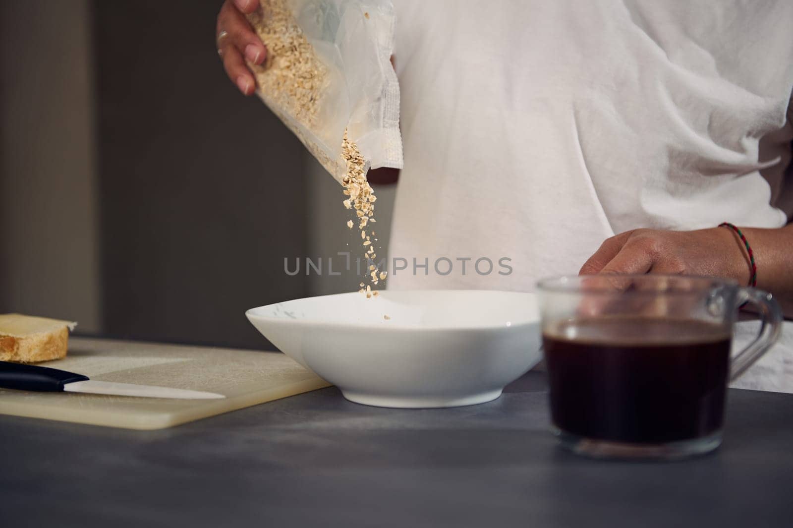 Close-up woman pouring oat flakes into a white bowl, preparing dry breakfast, standing t kitchen counter at home. Food and drink concept. Healthy eating