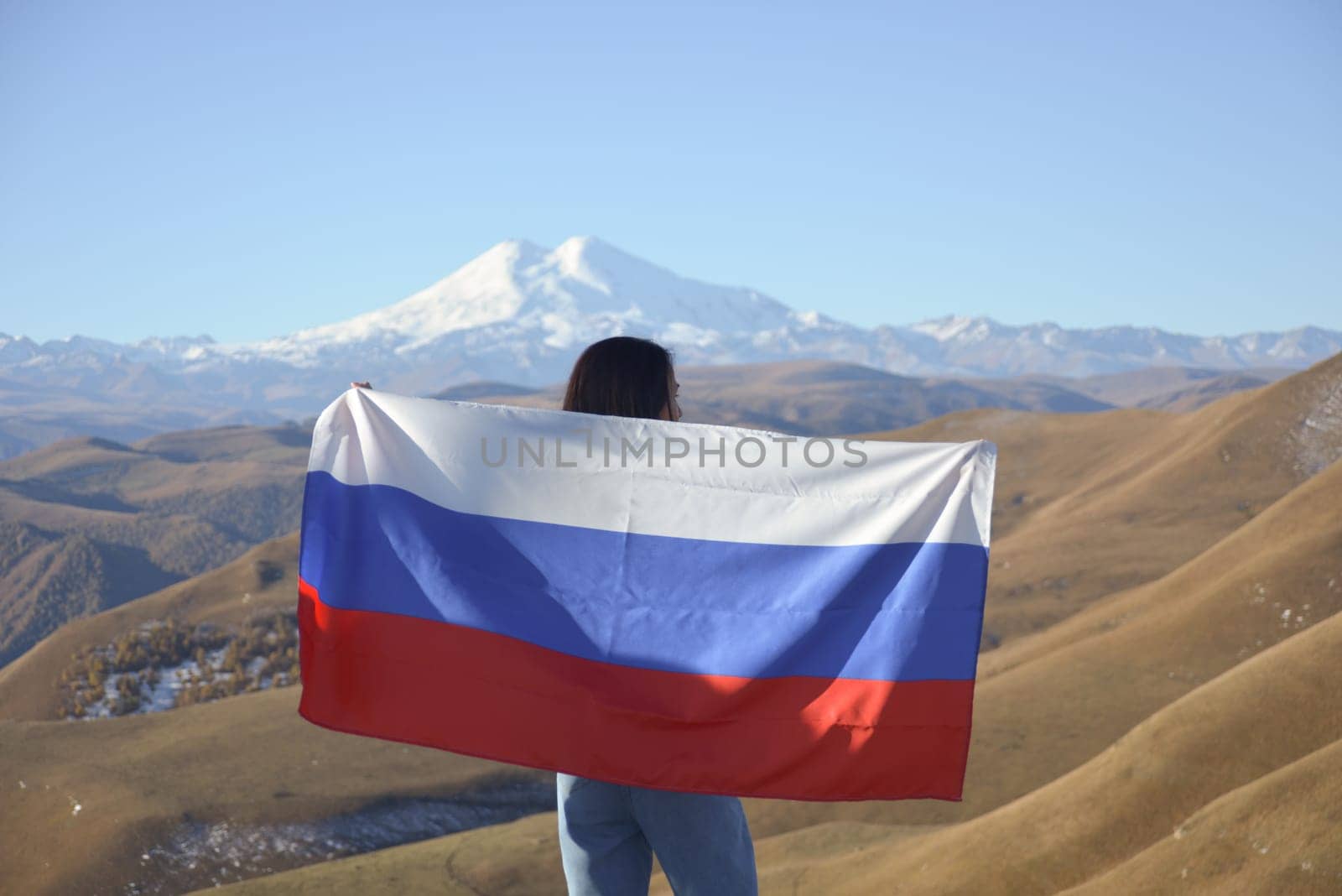 A young brunette woman stands against the backdrop of the snow-capped Mount Elbrus, looking at the beauty of the mountains, holding a Russian flag in her hands. Tricolor against the backdrop of snow-capped Mount Elbrus.