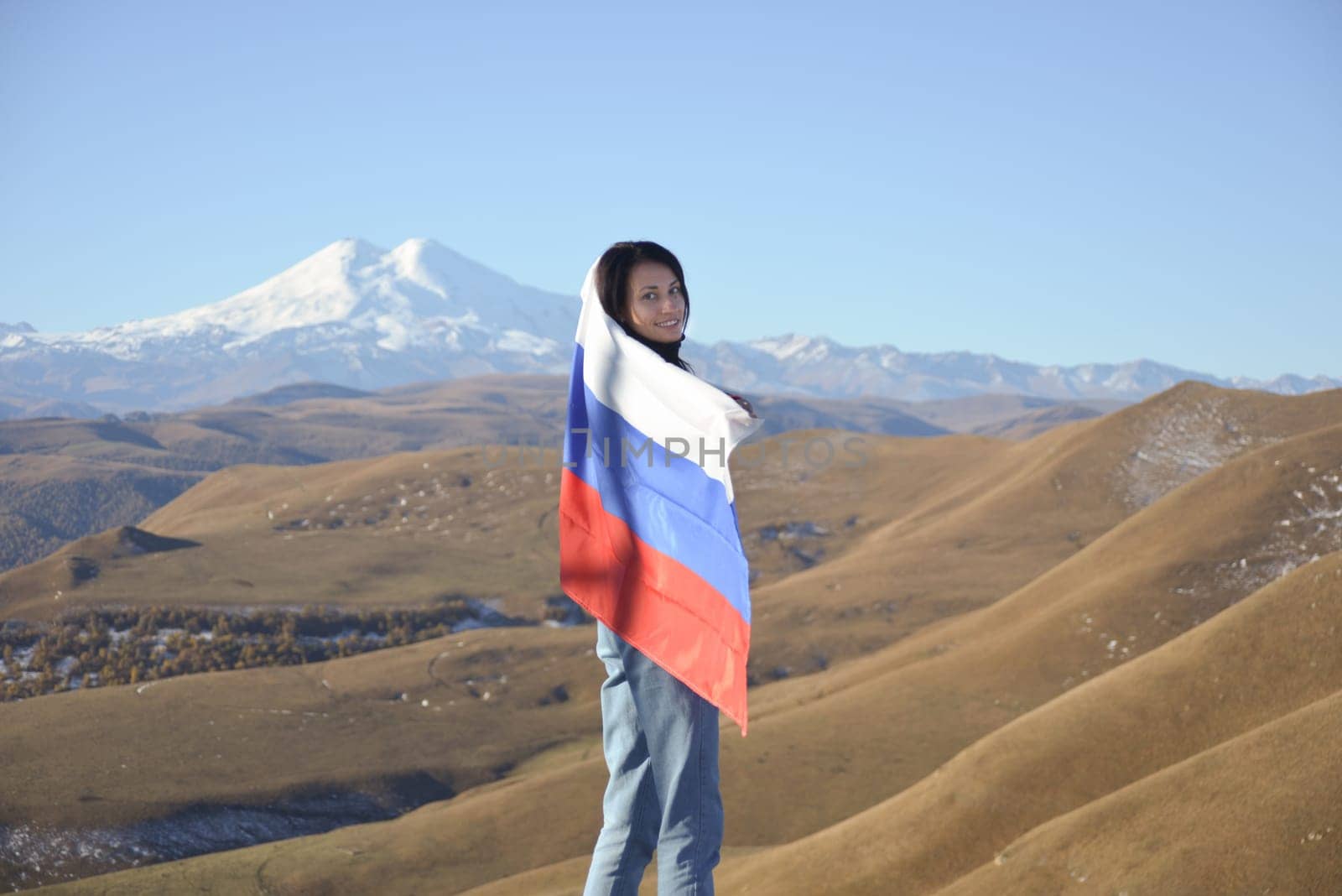 A young brunette woman stands against the backdrop of the snow-capped Mount Elbrus, looking at the camera, a Russian flag covers her shoulders. Tricolor against the backdrop of snow-capped Mount Elbrus by Ekaterina34