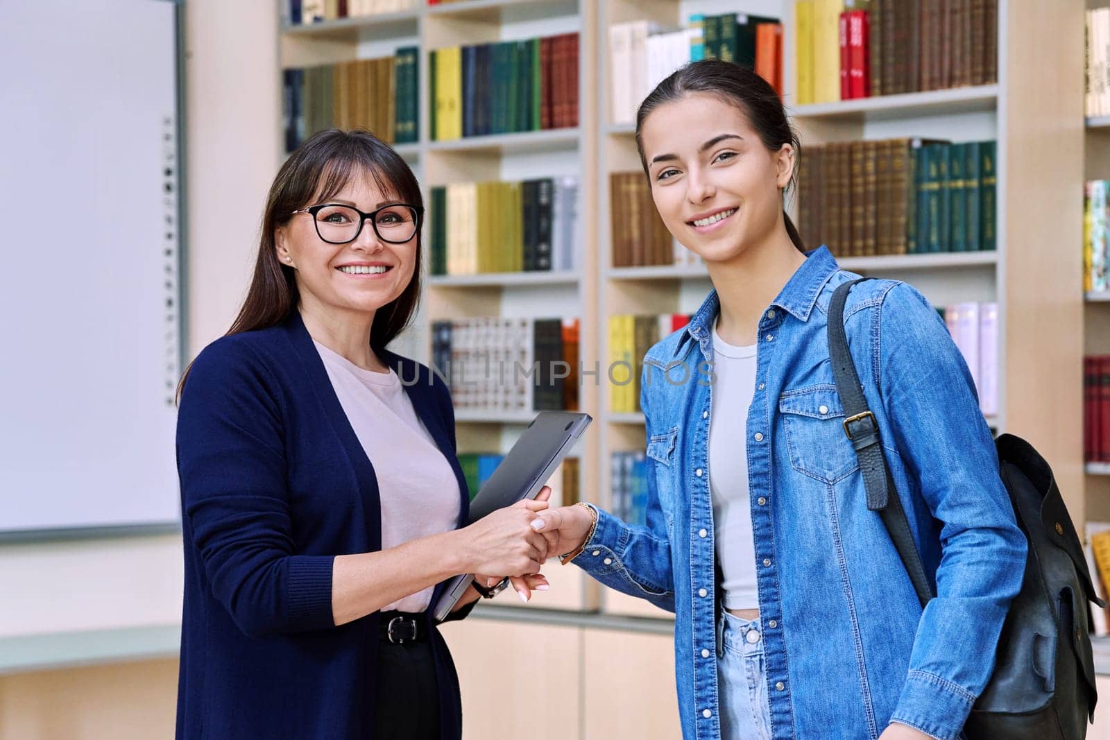 Female teacher talking to teenage girl high school student, shaking hands with each other, inside educational building library office. Education, training, teaching, adolescence youth concept