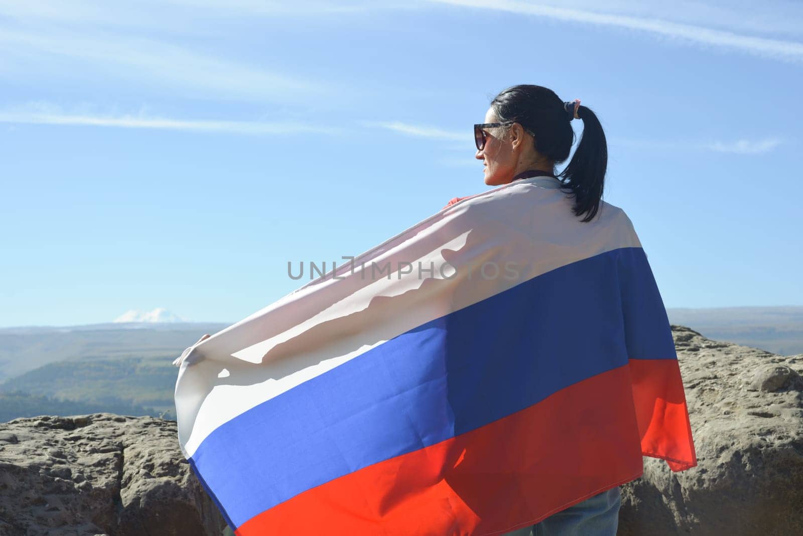 A girl tourist with a Russian flag on her shoulders stands on the top of a mountain and enjoys the beautiful views of the Caucasus Mountains