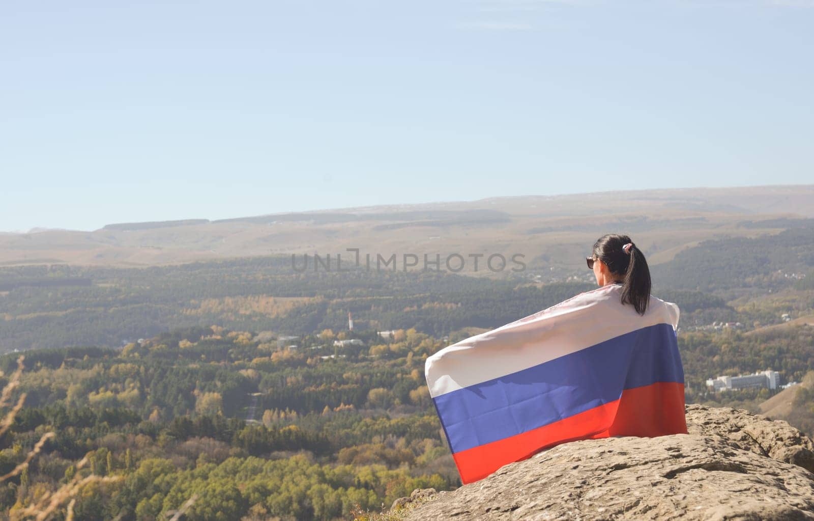 A young brunette woman sits on a stone and looks at the mountains around, her shoulders are covered with a Russian flag.