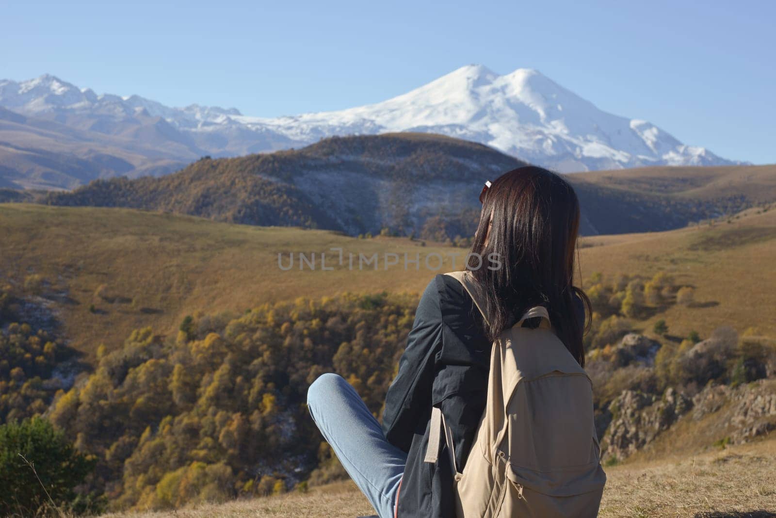 A female traveler with a backpack behind her sits on the grass and enjoys the views of Elbrus. View of Elbrus, North Caucasus, Russia. by Ekaterina34