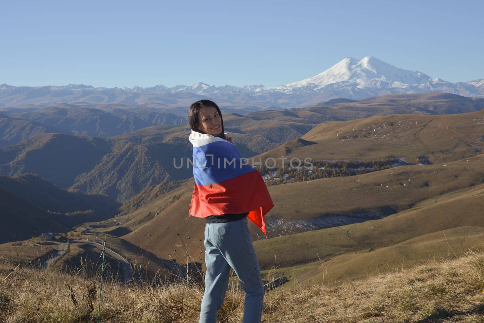 A young brunette woman stands against the backdrop of the snow-capped Mount Elbrus, looking at the camera, a Russian flag covers her shoulders. Tricolor against the backdrop of snow-capped Mount Elbrus.