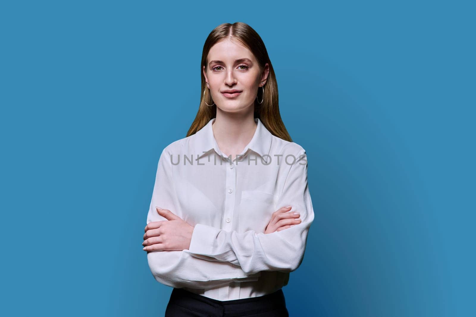 Portrait of young teenage smiling female in white shirt with crossed arms on blue studio background. Confident beautiful happy college student girl looking at camera