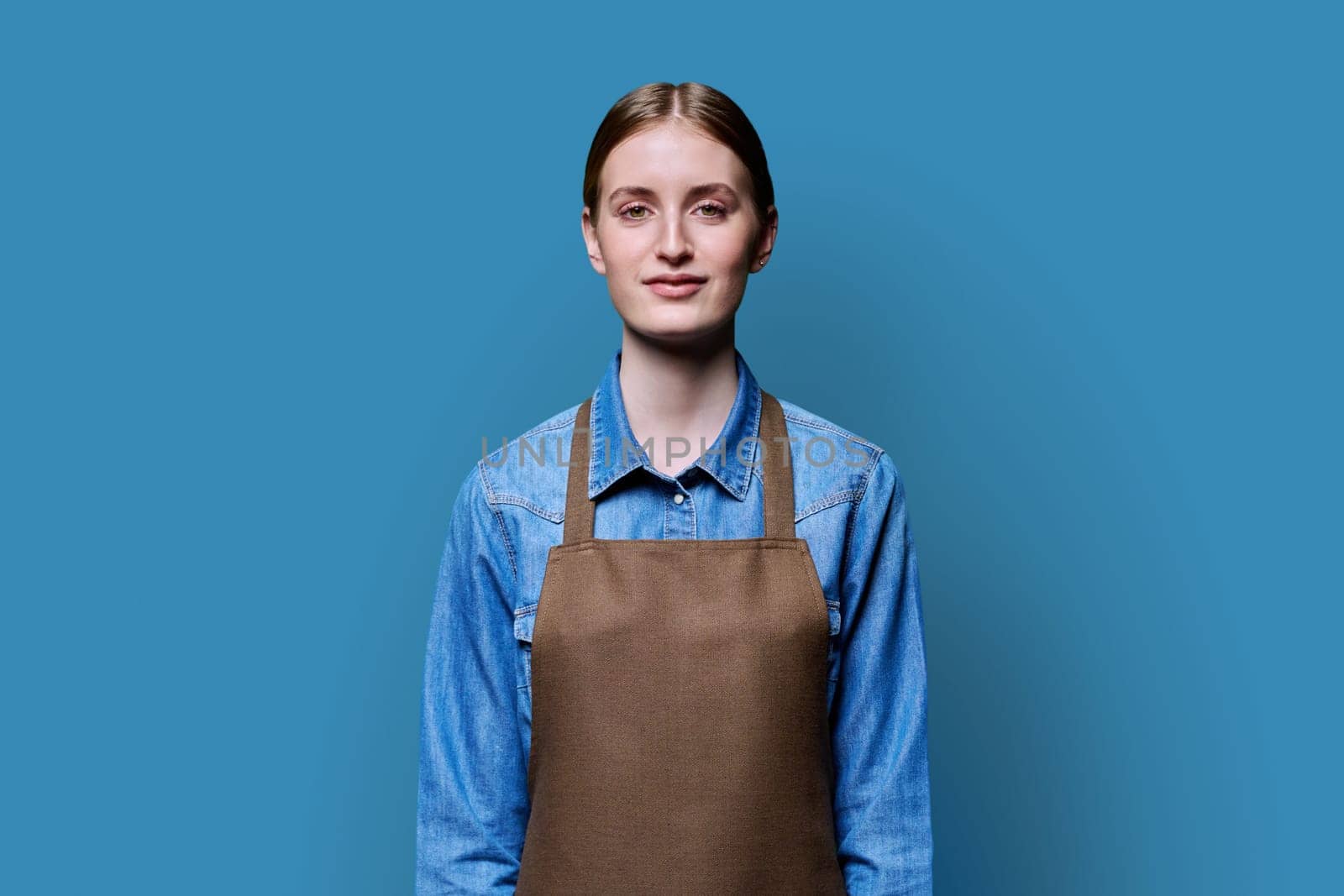 Portrait of young smiling confident woman in apron on blue studio background. Successful positive female looking at camera. Worker, startup, small business, service sector, staff, youth concept