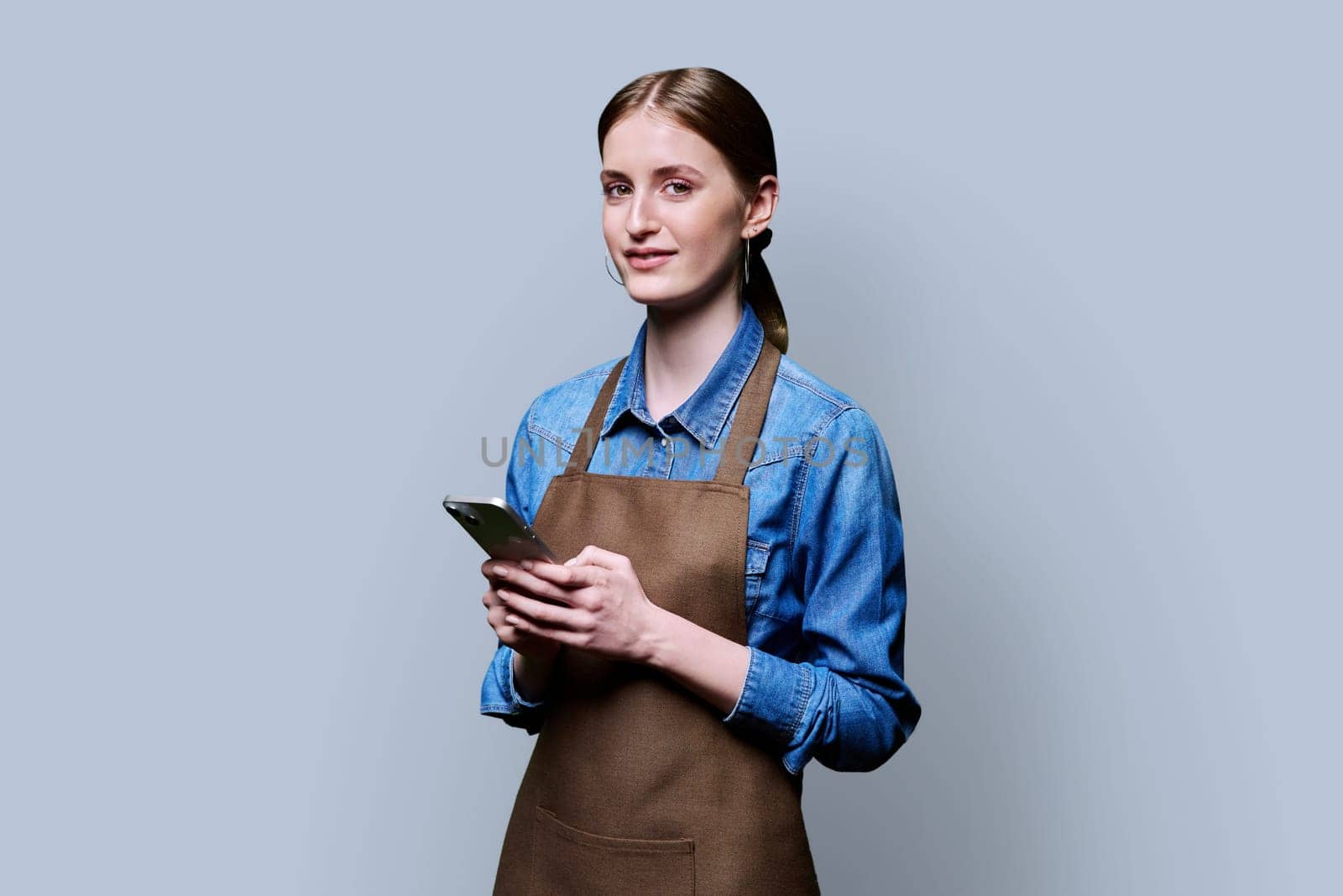 Young female worker in an apron looking at camera holding smartphone on gray studio background. Work, business, online internet services, mobile apps applications, technology concept
