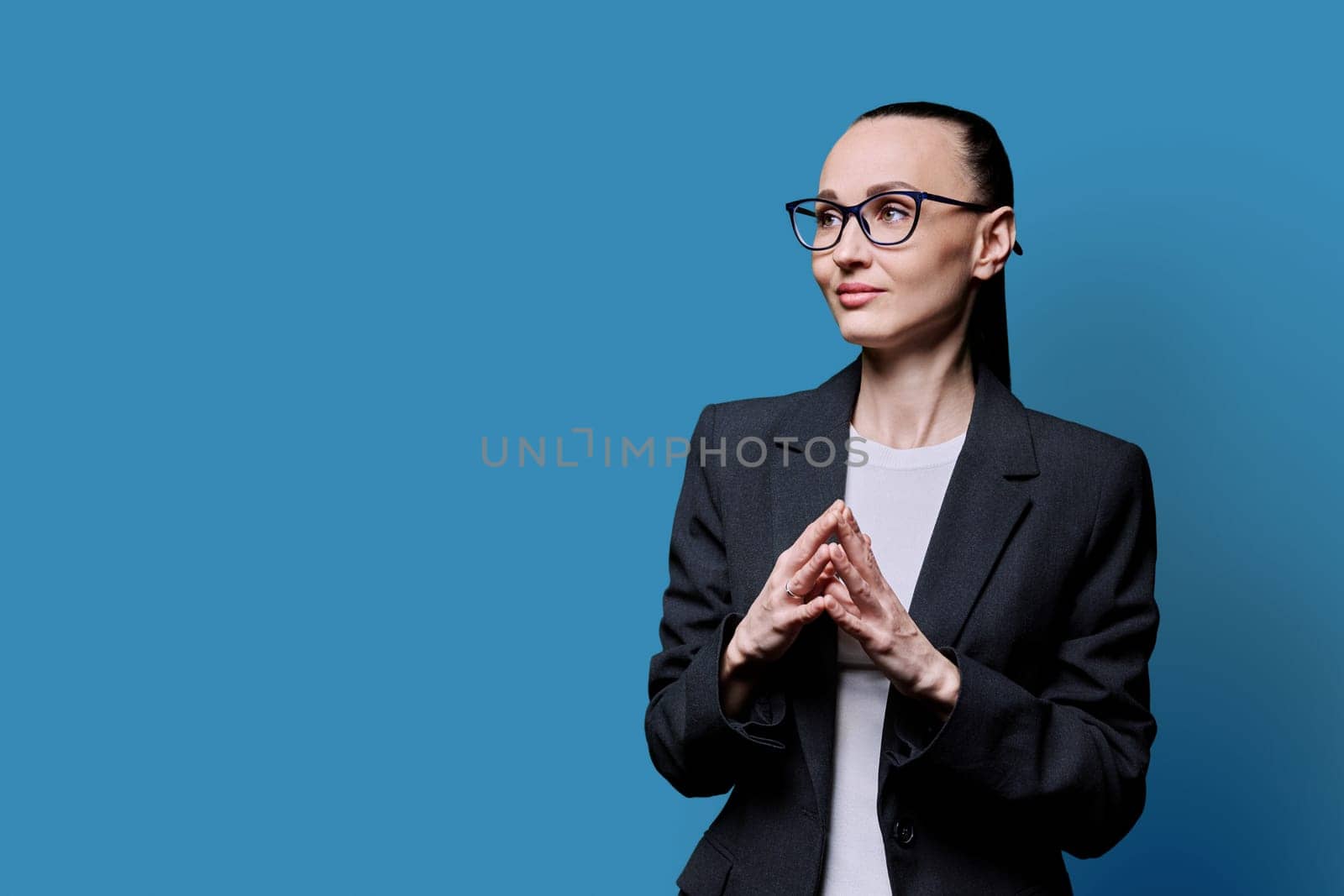 Portrait of thinking serious 30s business woman on blue studio background. Confident female in glasses suit looking to side at space for image text copy space. Business work teaching job career people