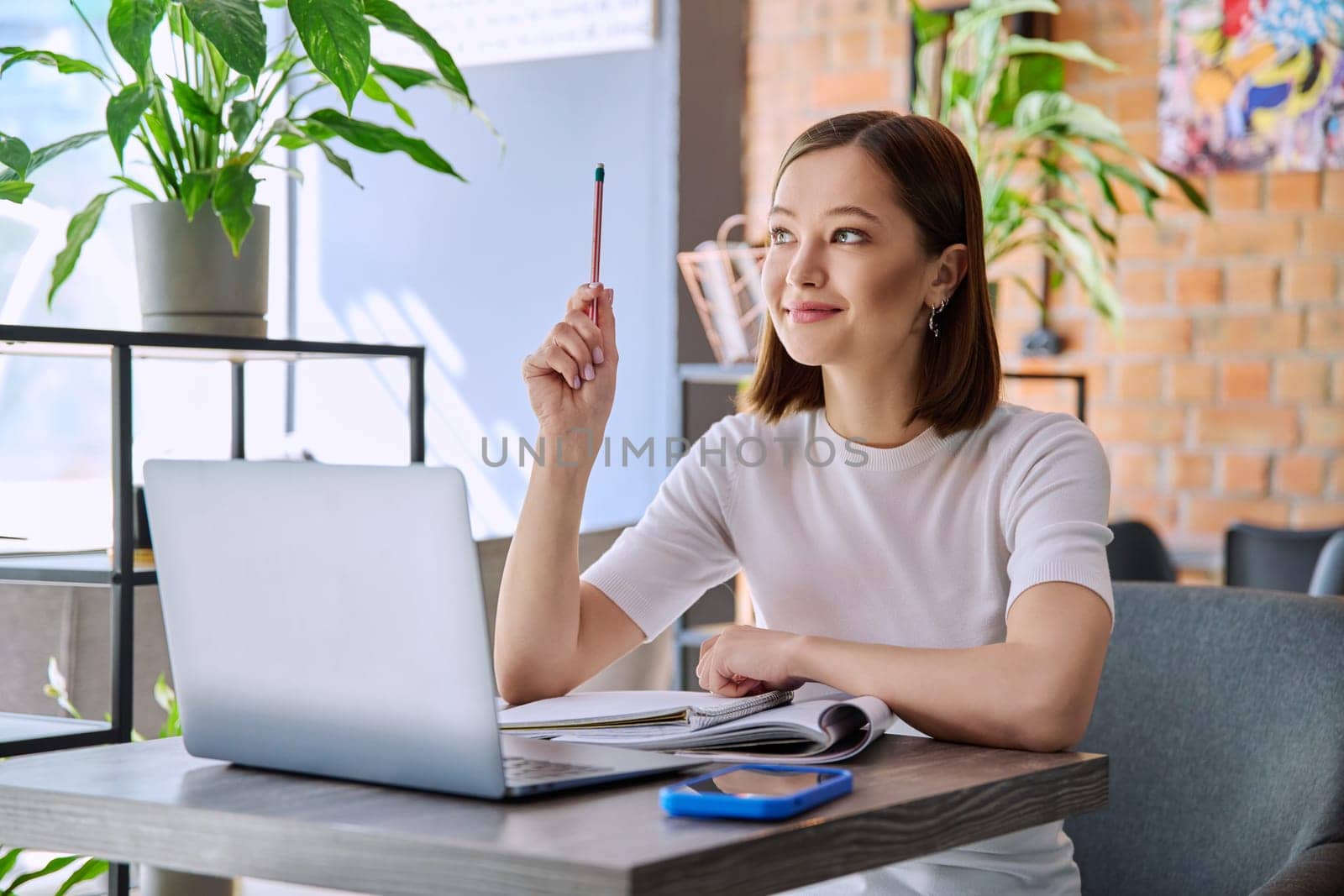 Young female college student studying using laptop, writing in notebook while sitting in coworking cafe. E-learning, education, lifestyle, youth concept