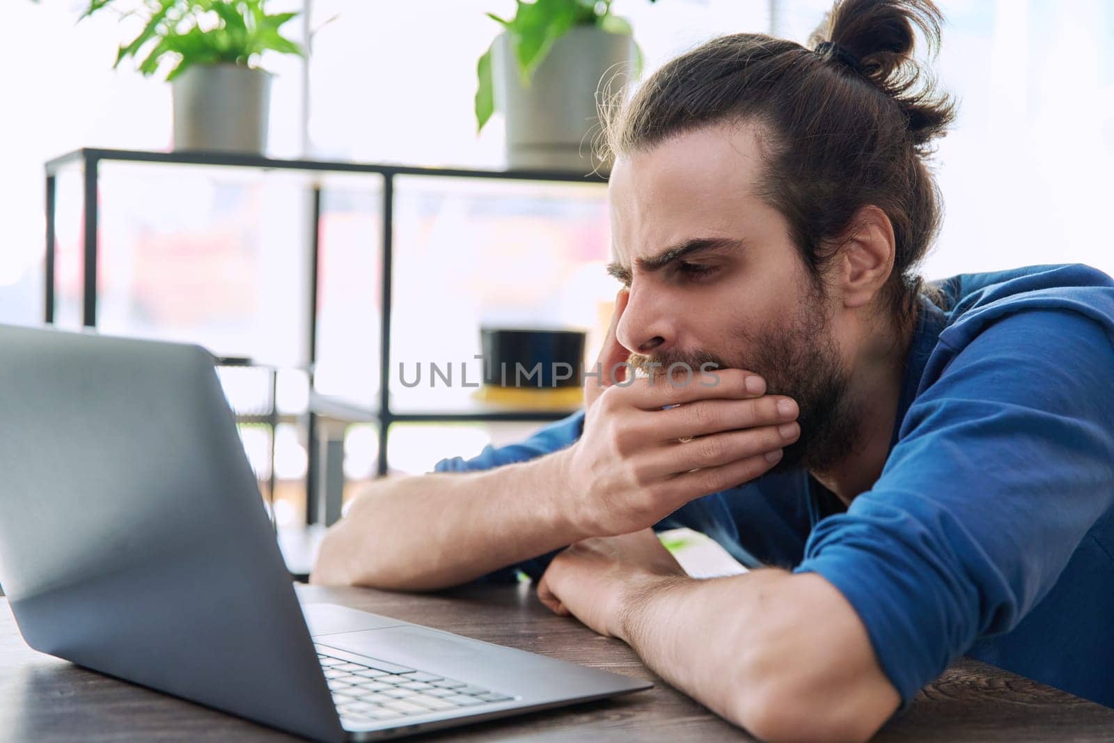 Young tired sleepy man looking at laptop computer, yawning covering his mouth with hand. Fatigue, drowsiness, lifestyle concept