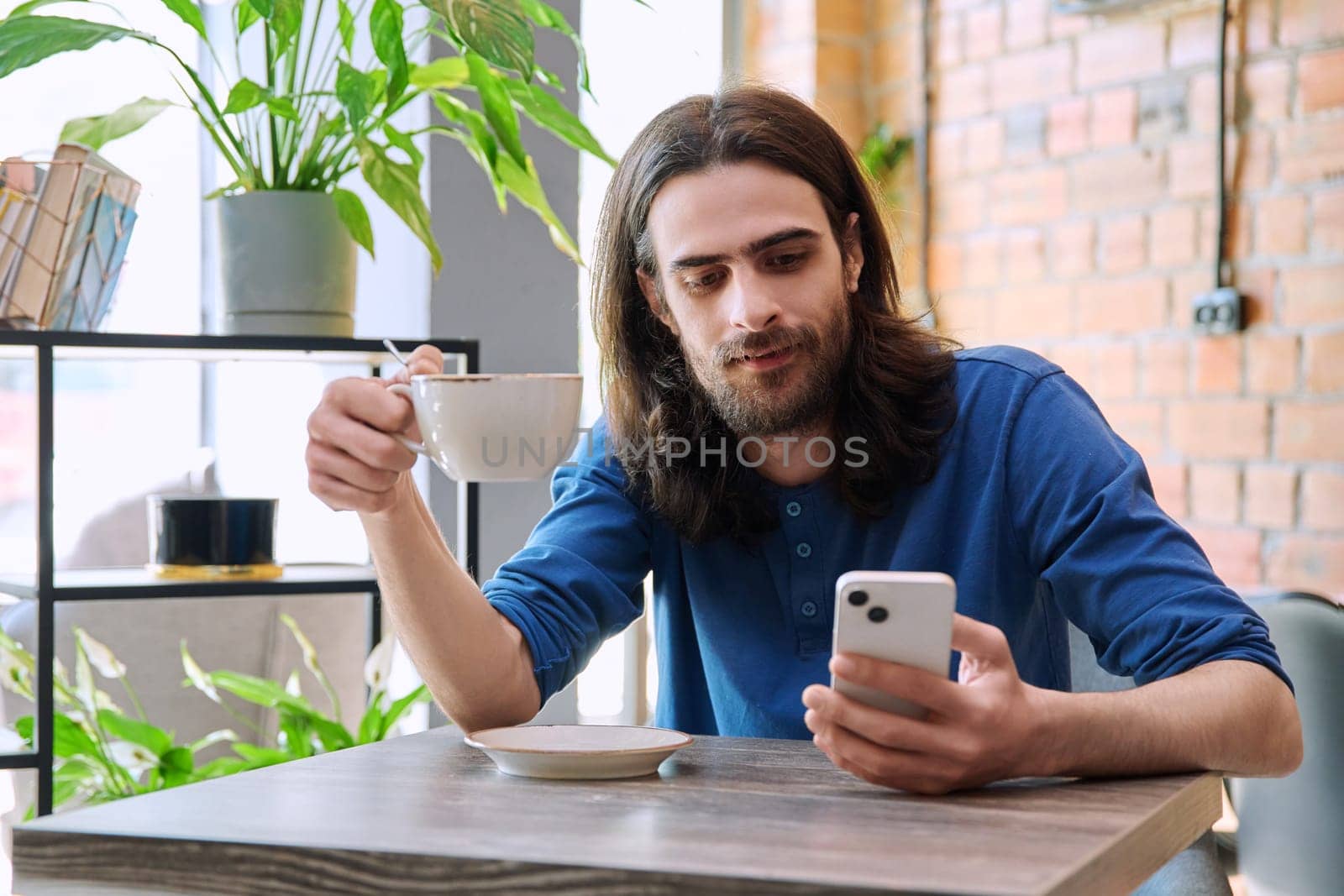 Young 30s stylish handsome bearded with long hair man using smartphone, drinking cup of coffee, sitting in cafe. Mobile technologies Internet apps applications for leisure work business communication