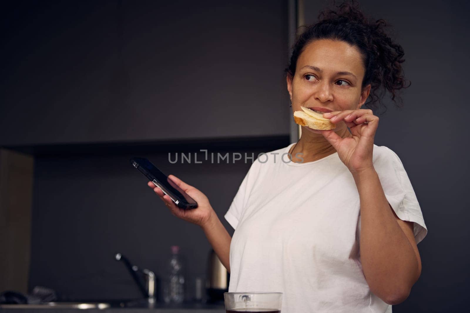 Authentic multi ethnic young adult woman 40s, eating sandwich with cheese and holding her smartphone in hands, smiling looking aside, standing at kitchen counter in minimalist home kitchen interior.