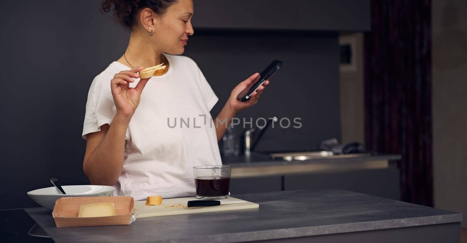 Gorgeous multi ethnic woman 40s, using smartphone, checking social media content, browsing web, scrolling newsfeed, standing at kitchen counter with a sandwich in hand, enjoying her breakfast at home