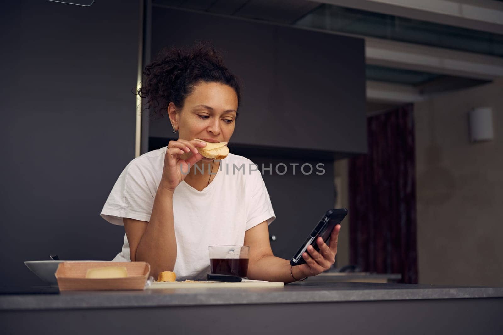 Beautiful multi ethnic woman 40s, eating sandwich with cheese and checking social media content on his smartphone, standing at kitchen counter in minimalist home kitchen interior.