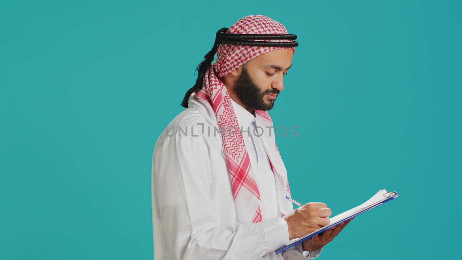 Arab man writing letter on papers standing over blue background, creating new schedule and taking notes. Young adult in muslim clothing looking at checklist for paperwork.