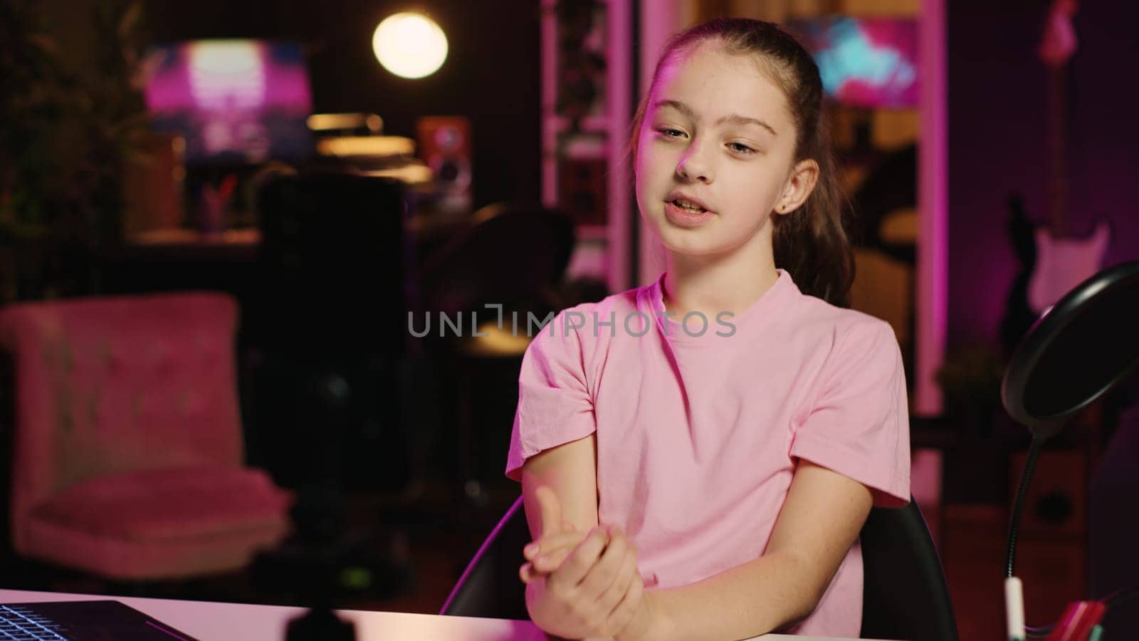 Charming young girl shooting video for social media platform in living room, talking about her day. Kid entertaining followers, doing content creation in neon lit apartment using smartphone on tripod