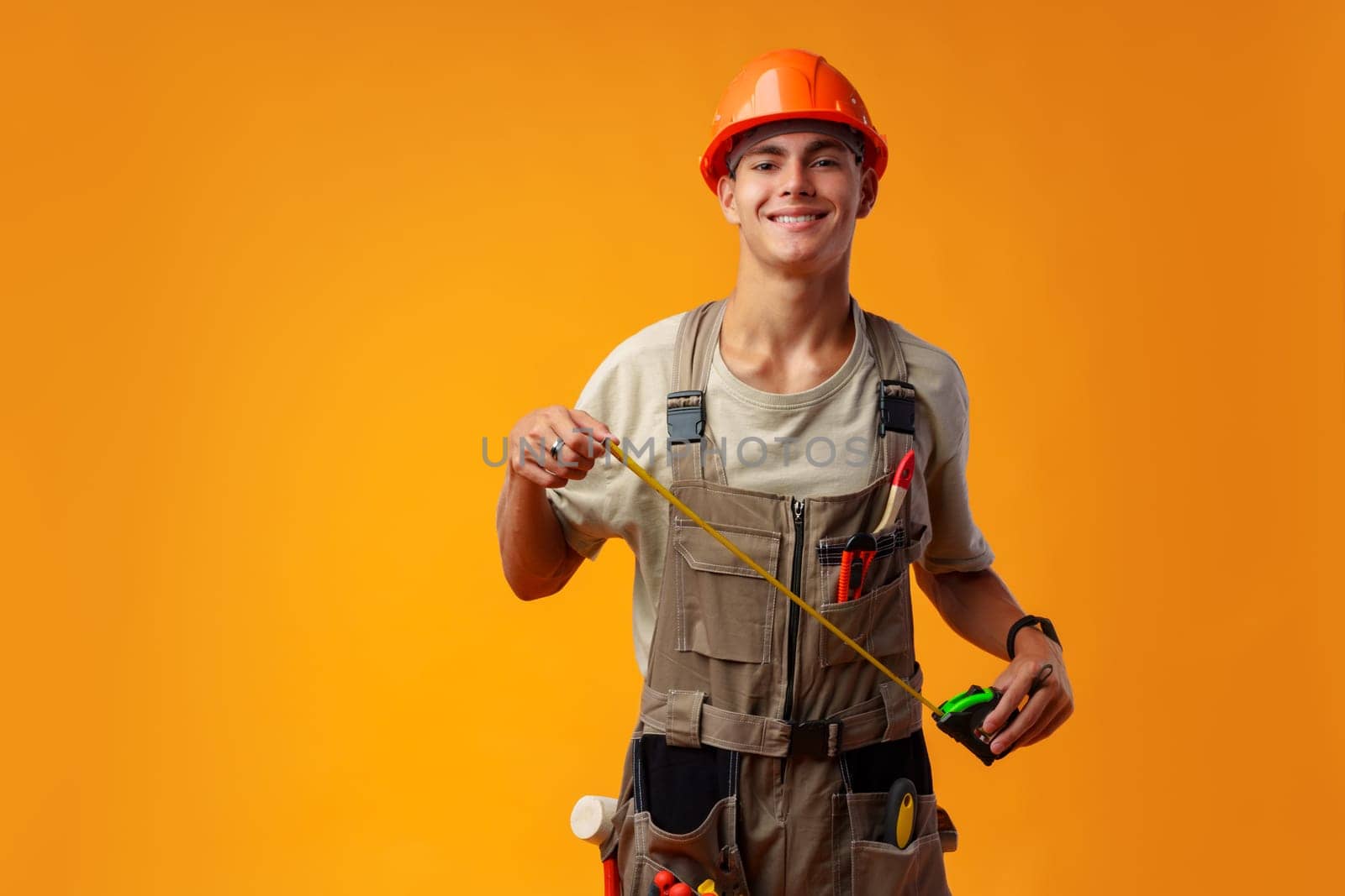 Young male builder holding measuring roulette against yellow background in studio