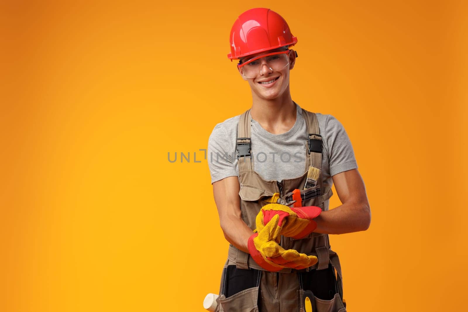 Young construction worker in helmet and uniform posing on yellow background in studio, close up