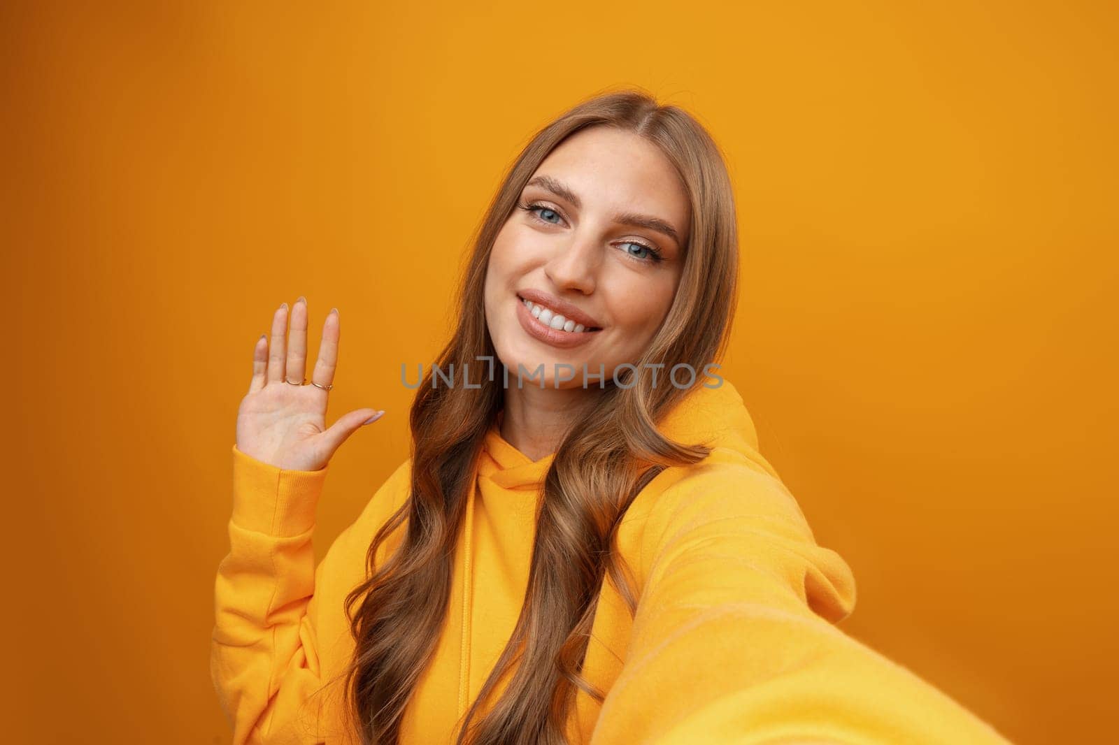 Happy cheerful woman with curly hair waving hello at smartphone camera on yellow background close up