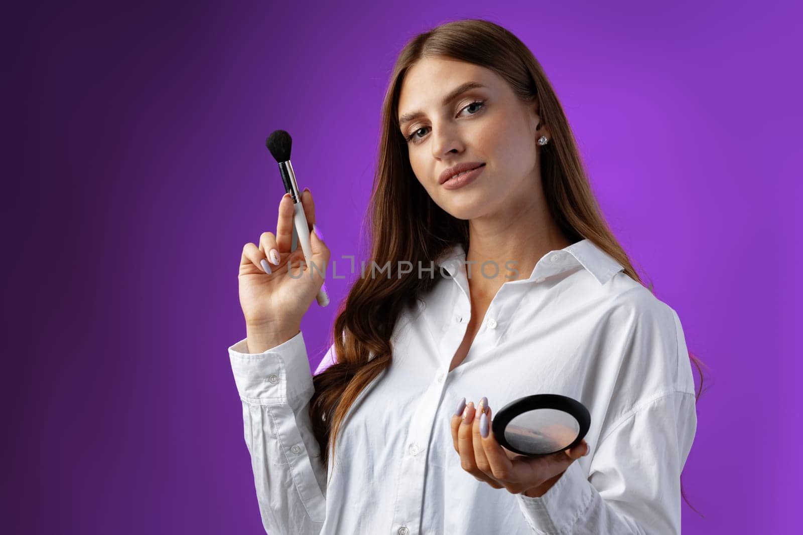 Young woman with makeup holding powder brush against purple background in studio