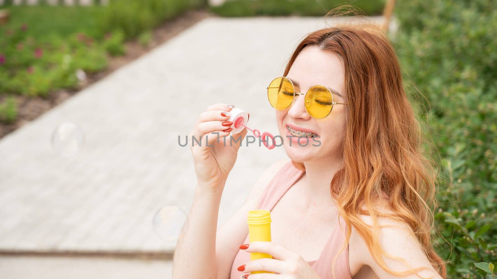Young red-haired woman blowing soap bubbles outdoors. Girl with braces on her teeth