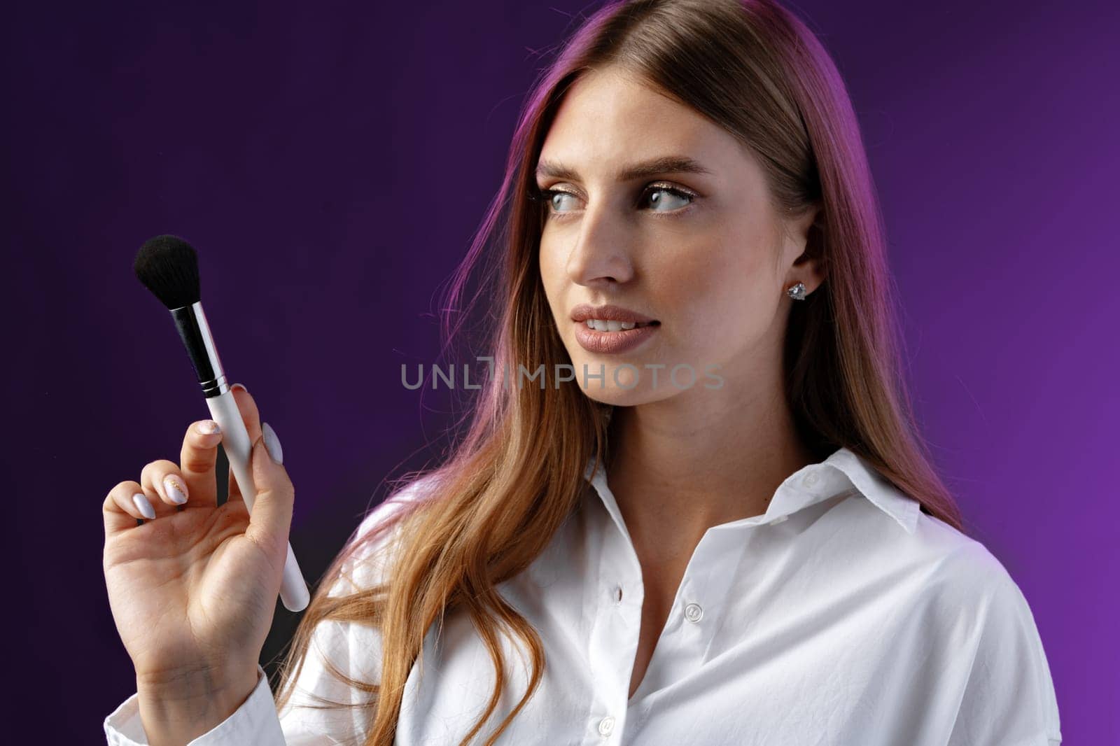 Young woman with makeup holding powder brush against purple background in studio