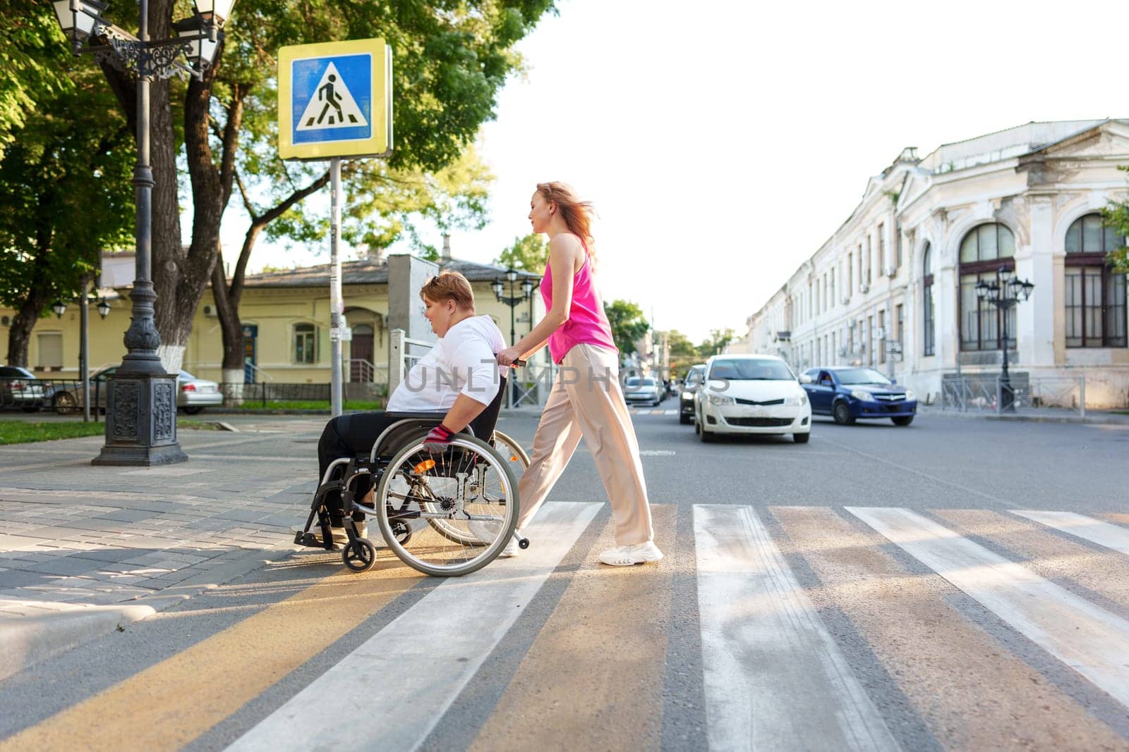 Young female caregiver pushing wheelchair with mature female person with disability across city street