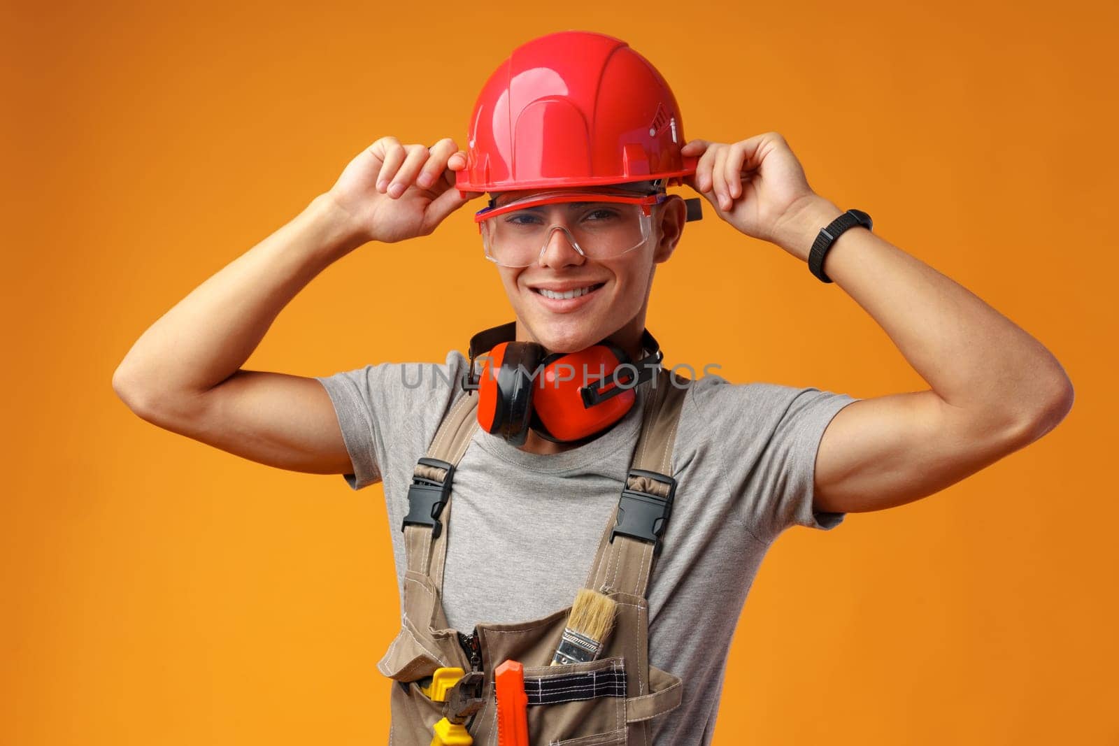 Young construction worker in helmet and uniform posing on yellow background in studio, close up