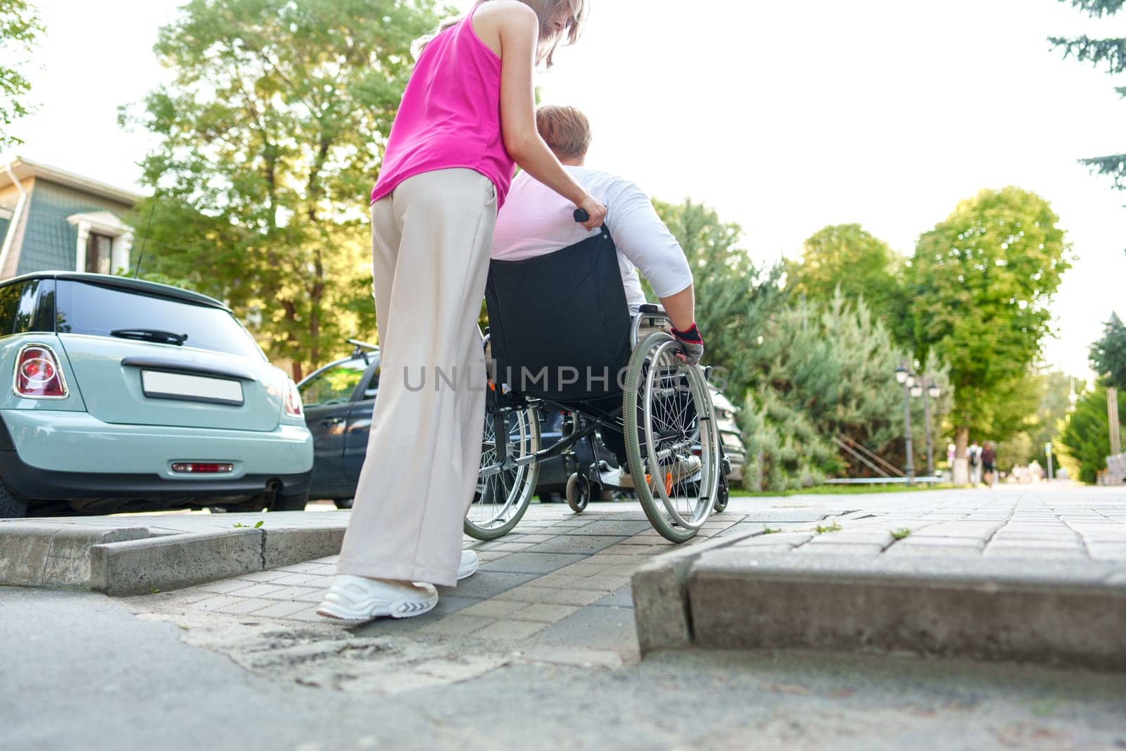 Young female caregiver pushing wheelchair with mature female person with disability across city street